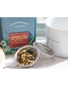 Dark blue tin with metal clasp of Cartwright & Butler green tea with jasmine blend with colourful illustrations of flowers, leaves, and a bird. With a small amount of tea blend placed in front of the tin