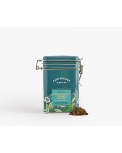 Dark blue tin with metal clasp of Cartwright & Butler peppermint tisane blend with colourful illustrations of flowers, leaves, and a bird. With a small amount of tea blend placed in front of the tin