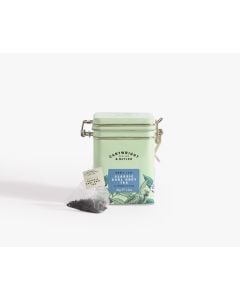 Light green tin with metal clasp of Cartwright & Butler classic earl grey tea with colourful illustrations of flowers, leaves, and a bird. With a tea bag placed in front of the tin